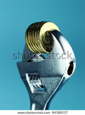A stack of Gold Dollar Coins being squeezed in a silver adjustable spanner, against a light pastel blue background and asking the questions how tight  is you household budget?