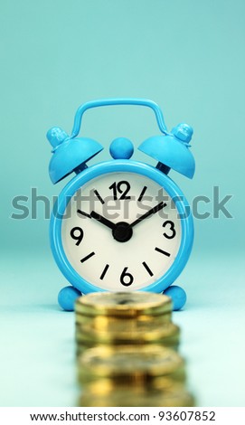 A light blue alarm clock placed in front  four stacks of gold coins with pastel blue background, hence the phrase, time is money.