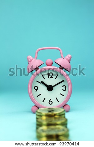 A pastel pink alarm clock placed in front  stacks of gold coins with pastel blue background, hence the phrase, time is money.