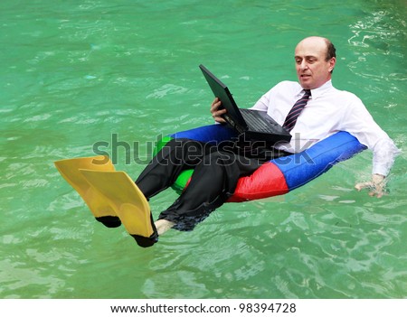 A businessman working from home dressed in business attire floating in a swimming pool on his laptop and wearing yellow flippers, and testing the water with his left hand