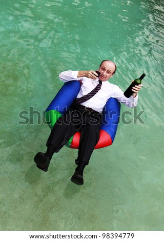 A business man dressed in business attire in a swimming pool, floating with a glass of red wine in one hand and the bottle in the other.