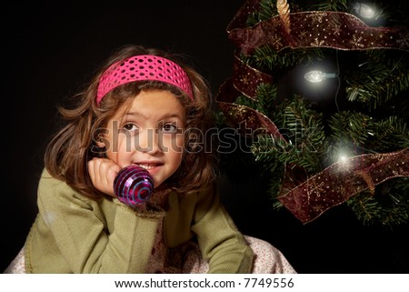 A girl thinking nearby a christmas tree holding a christmas ball.