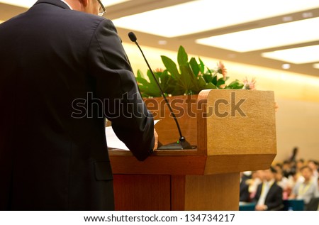 Business man is making a speech in front of a big audience at a conference hall.