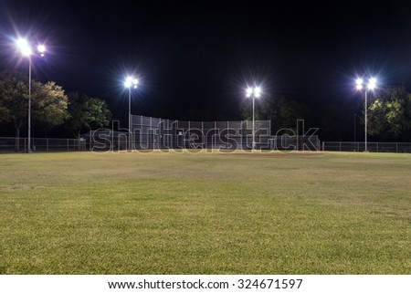 Night photo of an empty baseball field at night looking back toward home plate from right field with the lights on and contrasting against the blackness of the night sky.