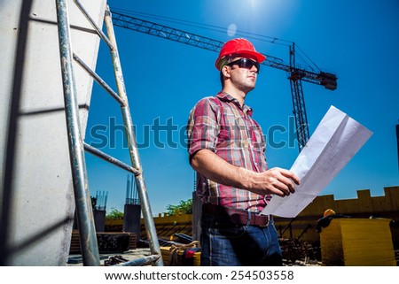 Real Engineer looking at his blueprint plan on construction site, ladder and crane seen in the background