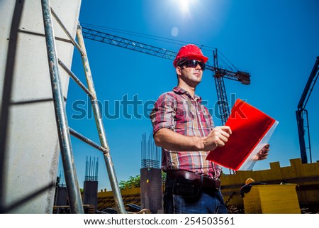 Real Engineer looking at his blueprint plan on construction site, ladder and crane seen in the background