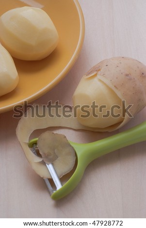 Peeling potato with peeler, two peeled potato on yellow plate, small group of objects on wooden background.