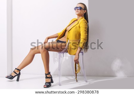 Full-length portrait young elegant woman in the yellow jacket, shorts and shoes with heels on a transparent chair with banana. Fashion studio shot.