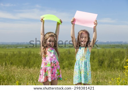Two baby girls in summer dresses on the nature of holding the colored sheets of paper. Summer, sun, green, meadow.