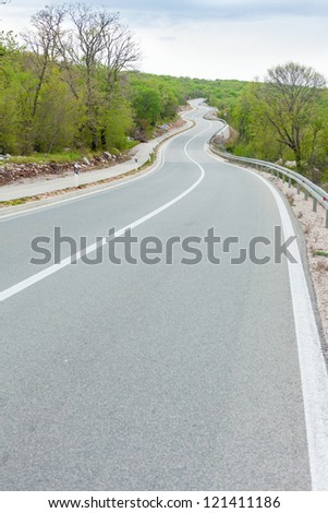 Curving black asphalt road with white marking lines from low point at roadside, trees, clouds on blue sky in background.