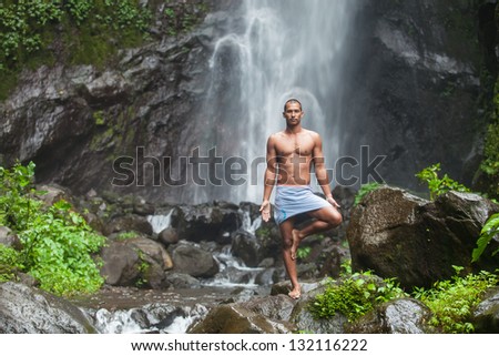 Young handsome man enjoying yoga at waterfall in the tropics