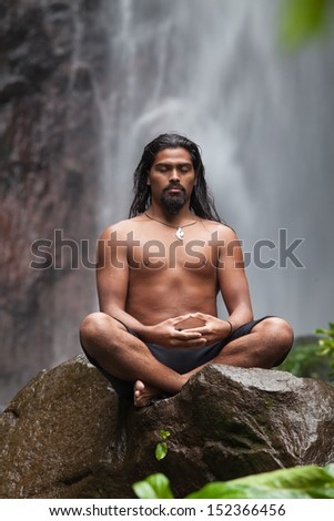 Man sitting in meditation on rock at waterfall in tropical rainforest