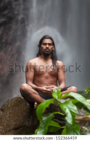 Man sitting in meditation on rock at waterfall in tropical rainforest