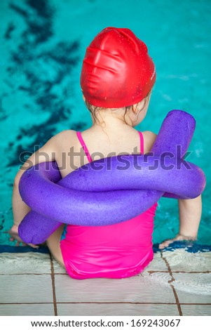 Little girl in the pink swimwear and red cap sitting on the edge of pool