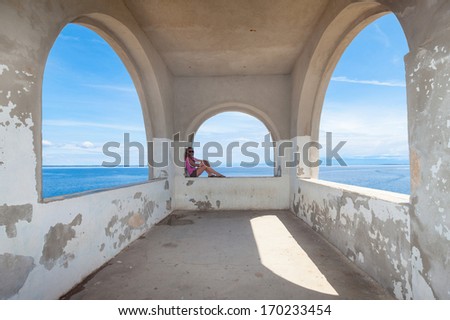 Young woman in the pink shirt sitting on the arch shaped stone window by the sea.