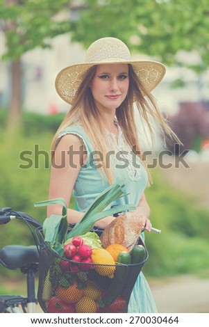 Attractive blonde woman with straw hat standing in the park and posing next to bike with basket full of groceries. Post processed with vintage filter.