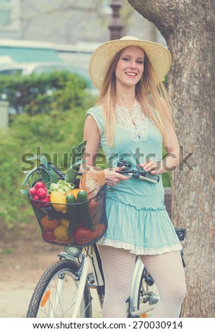 Attractive blonde woman with straw hat standing in the park and posing next to bike with basket full of groceries. Post processed with vintage filter.