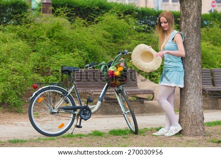 Attractive blonde womanwith straw hat standing in the park and posing next to bike with basket full of groceries.