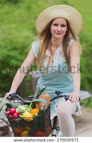 Attractive blonde woman with straw hat riding a bike with basket full of groceries.