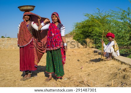 GODWAR REGION, INDIA - 14 FEBRUARY 2015: Rabari women stand in field wearing sarees and upper-arm bracelets with man sitting in background. Rabari are an Indian community in the state of Gujarat.