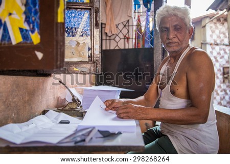 MUMBAI, INDIA - 16 JANUARY 2015: Elderly Indian man sits outdoors in front of home and punches holes into business paper.
