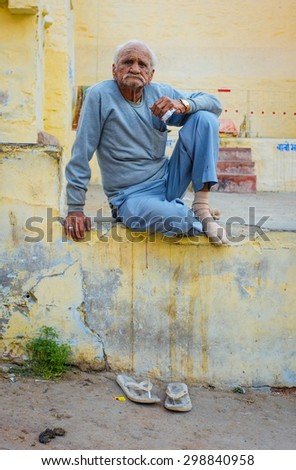 JODHPUR, INDIA - 07 FEBRUARY 2015: Elderly man sitting cross-legged on wall with slippers below him in dirty street. Common scene to see people hanging out on Jodhpur streets in late afternoon.
