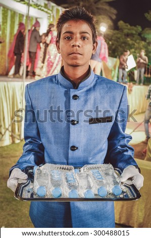 JODHPUR, INDIA - 08 FEBRUARY 2015: Young Indian boy wearing suit holds water bottles on tray working as waiter. Post-processed with grain, texture and colour effect.