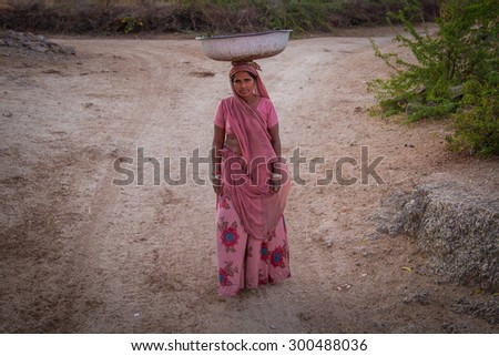GODWAR REGION, INDIA - 14 FEBRUARY 2015: Indian woman in pink sari stands alone in dirt track with metal container balanced on head.