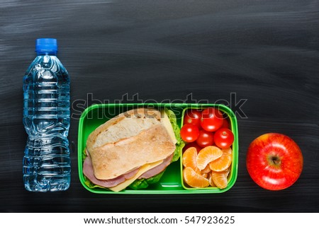 Sandwich, small tomatoes, tangerine, apple in plastic lunch box and bottle of water on black chalkboard.