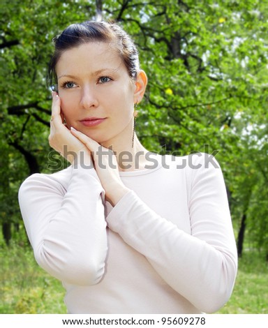 Close-up portrait of beautiful young girl in light-pink blouse at park with hands next to face