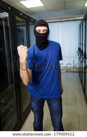 Masked cyber hacker wearing a balaclava threatens with fist in data center on the background.