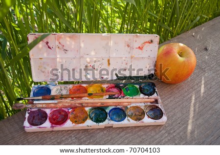 Water color paints, brushes and apple against a green grass