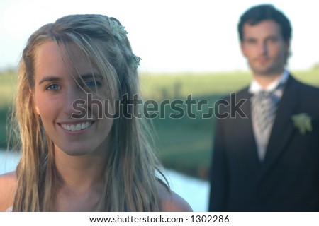 A bride and groom with the groom in the background. Lake-side country scene at sunset.