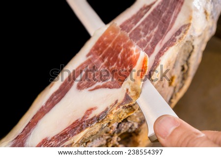 Knife with hand cutting serrano ham slices over black background, top view