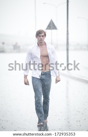 A Good looking male model walking in a road in the pouring rain with an open shirt and a denim jean and barefoot