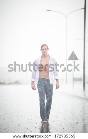 A Good looking male model walking in a road in the pouring rain with an open shirt and a denim jean and barefoot