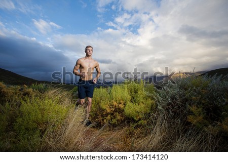 Male Fitness Model Running Allong A Trail In The Field, Wearing A Black Shirt And Shorts, With Big Clouds Ooverhead In The Sky.