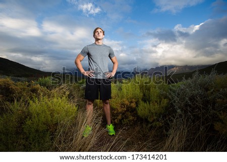 Male fitness model running along a trail in the field, wearing a black shirt and shorts, with big clouds overhead in the sky.