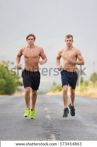 Two male models running on a tarmac road, bare chested and wearing shorts and running shoes.