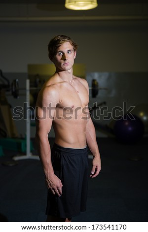 A Good looking male model flexing his muscles in a gym using weights, bare chest and wearing a short