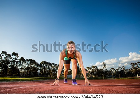 Fit female athlete standing / sitting at the start line to a sprint on a tartan athletics track with dramatic late afternoon lighting.