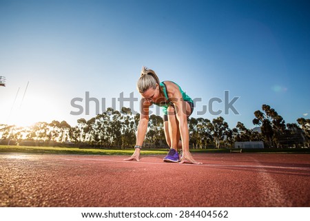 Fit female athlete standing / sitting at the start line to a sprint on a tartan athletics track with dramatic late afternoon lighting.