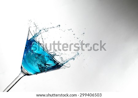 Close up image of a blue cocktail in a martini glass with an ice cube splashing into the liquid against a white background in studio