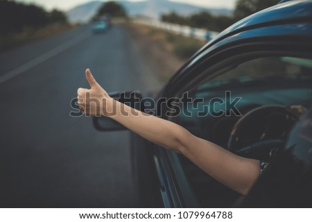 Young happy lady driving a car and making Like / Ok sign with hand out of window. Girl driving and enjoying road trip and travel, toned with soft autumn colors.