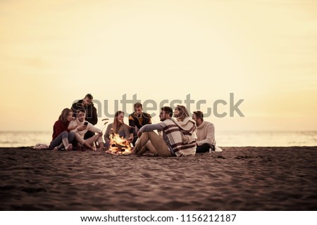 Group of young friends sitting by the fire at autumn beach, grilling sausages and drinking beer, talking and having fun filter