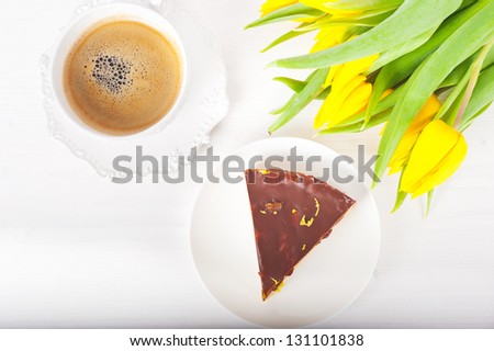 piece of layer cake with chocolate on a white plate, a cup of coffee and a bouquet of yellow tulips