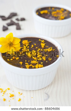 chocolate mousse in two tins of white, sprinkled with orange zest and decorated with a yellow flower on a wooden table, near the spill zest and slices of chocolate, vertical frame