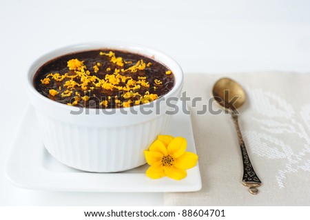 chocolate mousse in a white bowl on a white plate on a white linen napkin on the table, decorated with orange mousse orange zest, lies next to a yellow flower and a metal spoon