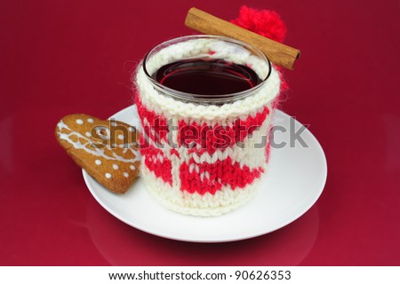 Mulled wine in a cup of knitted cover with a winter ornament, cinnamon stick, on a white saucer Christmas cookies in shape of heart on red background