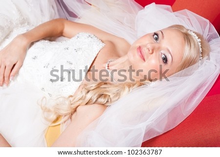 beautiful  bride wearing white wedding dress and veil lying on red sofa in interior of hall